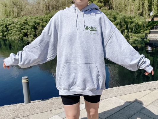Person wearing a gray hoodie with 'John' printed on it, standing by a body of water.